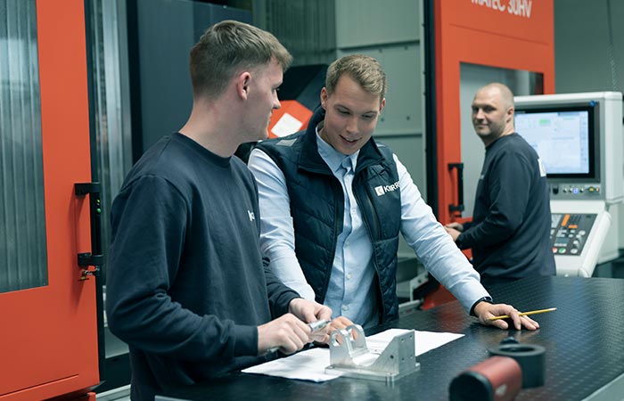 Zwei Mitarbeiter besprechen ein Metallteil während ein anderer Mitarbeiter die 5-Achs-Fräs-Maschine bedient