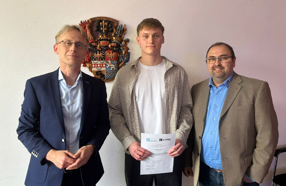 3 Männer vor einer weißen Wand, der Mann in der Mitte hält eine Bachelorarbeit.